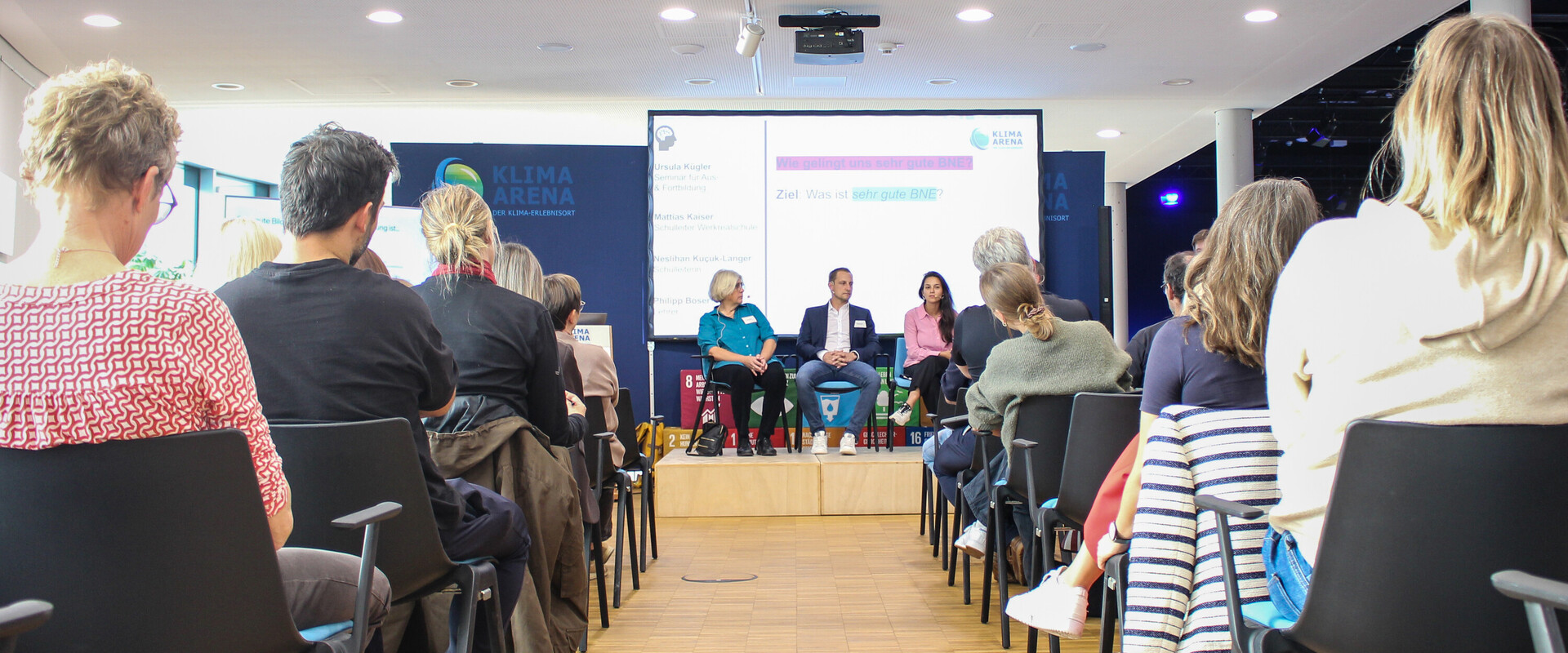 Blick durch die Stuhlreihen auf die G&auml;ste einer Podiumsdiskussion auf der B&uuml;hne